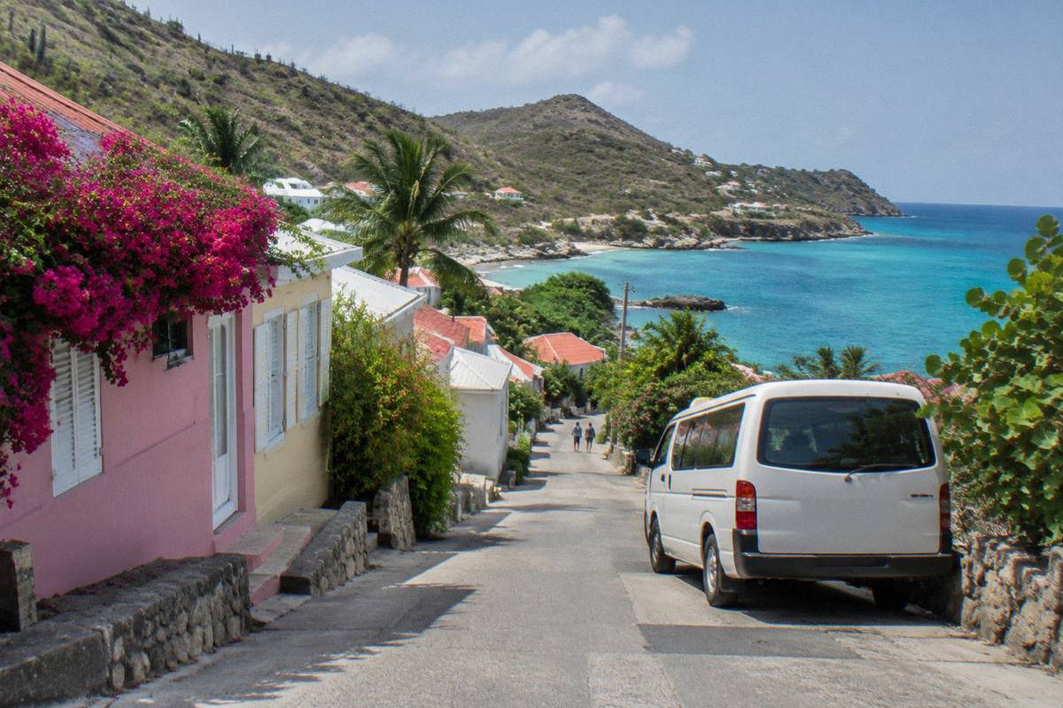 A white taxi van parked on a small street beside pastel-coloured shopfronts