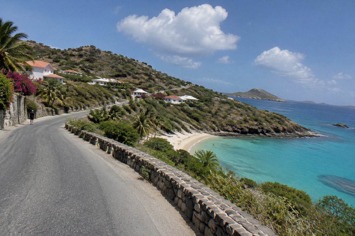 An open coastal road curving above a small sandy cove with calm water