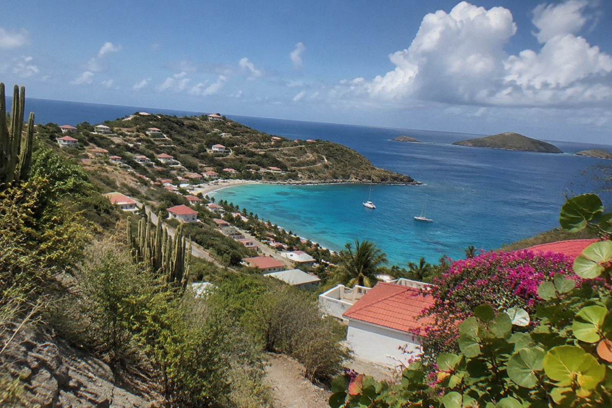 A hillside view over Saint Barthelemy with red rooftops, green slopes and open sea beyond.