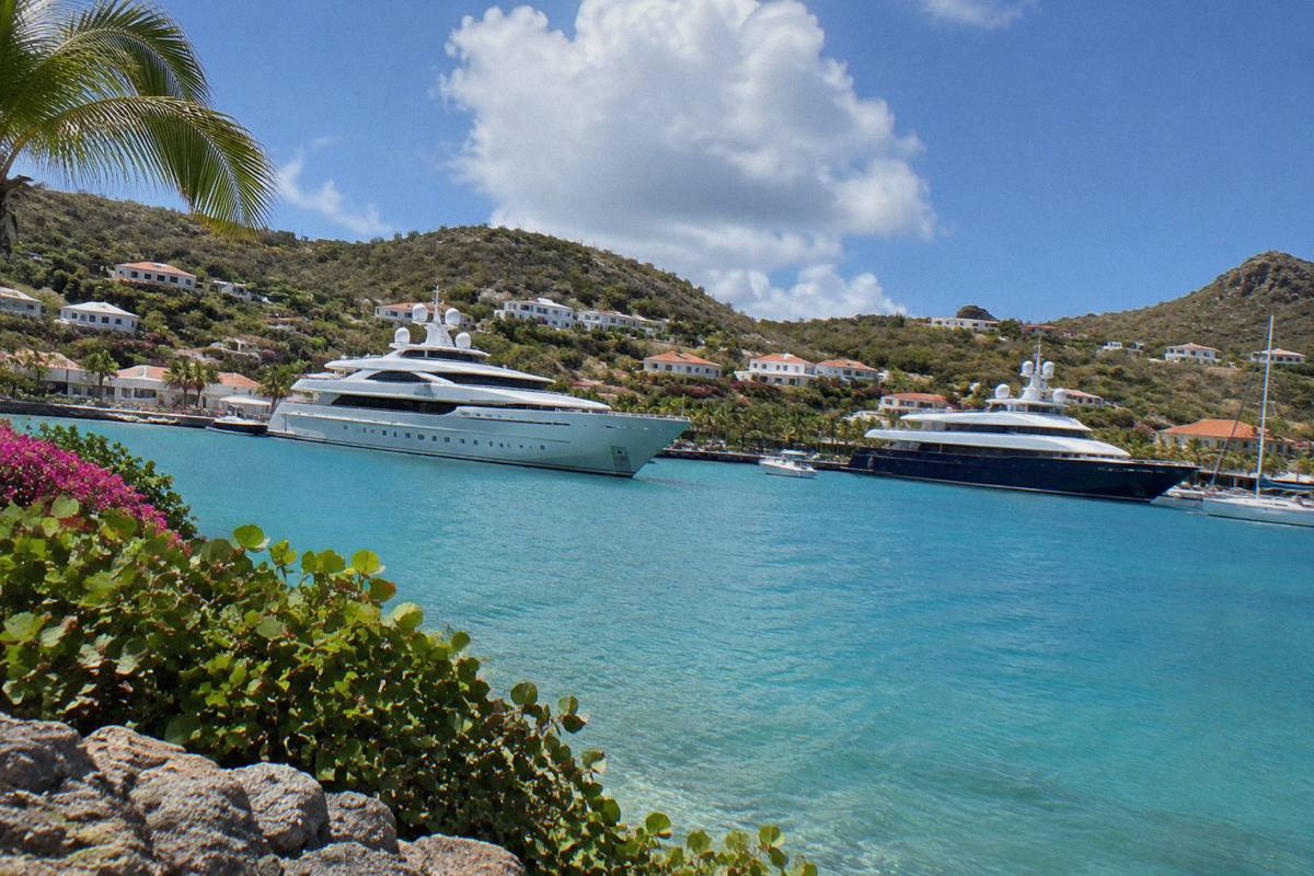 Superyachts moored in a harbour with hillsides of low buildings behind them.