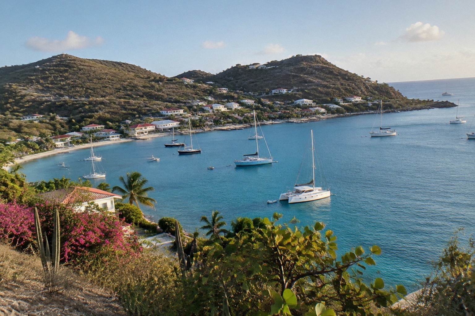 Yachts anchored in a small Caribbean harbour below hillsides with low white buildings at dusk.
