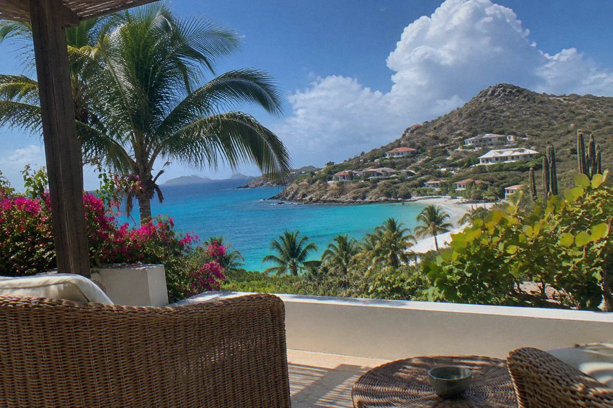 A hotel room terrace with rattan furniture and an ocean view through palm leaves
