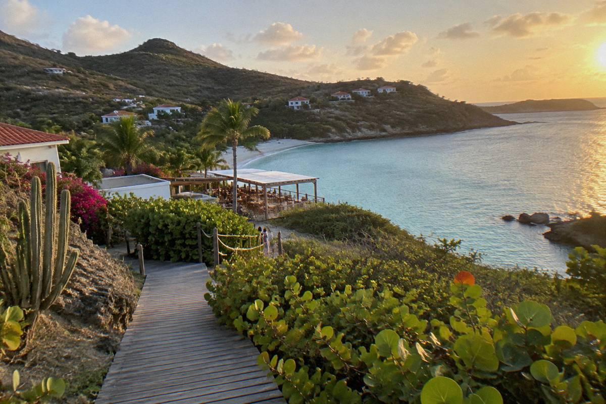 Wooden walkway crossing greenery toward a beachfront resort terrace beside a calm bay