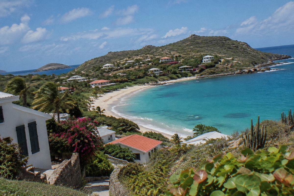 White resort buildings with dark shutters above a curving sandy bay on a green headland