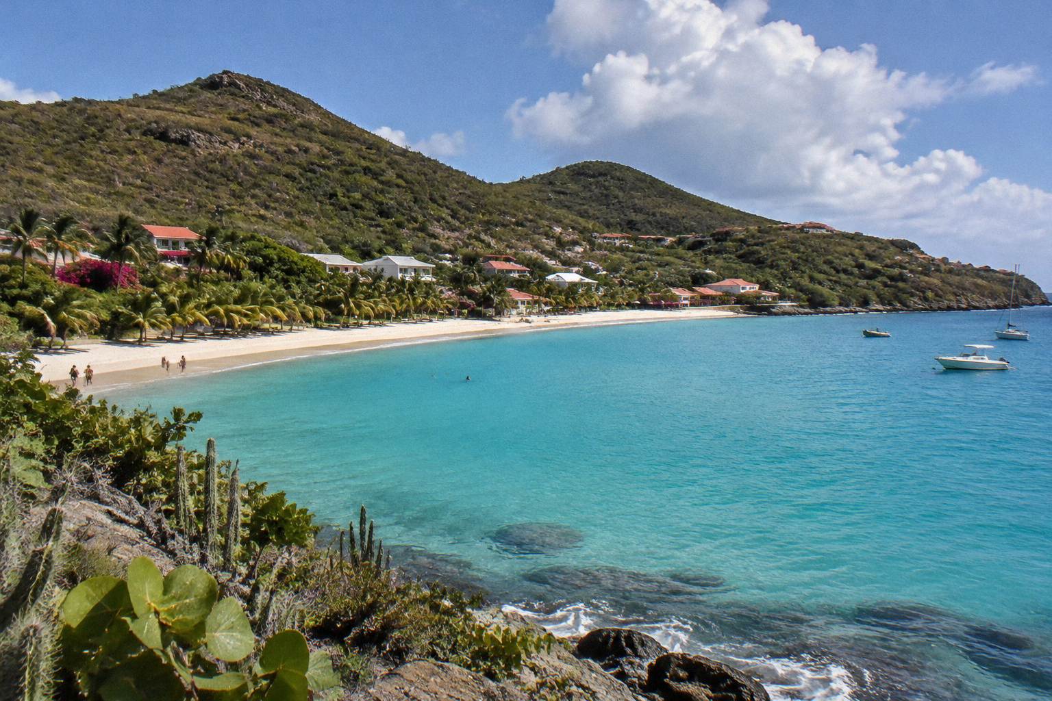 Calm turquoise bay with a palm-lined sandy beach and low green hills behind a small coastal village