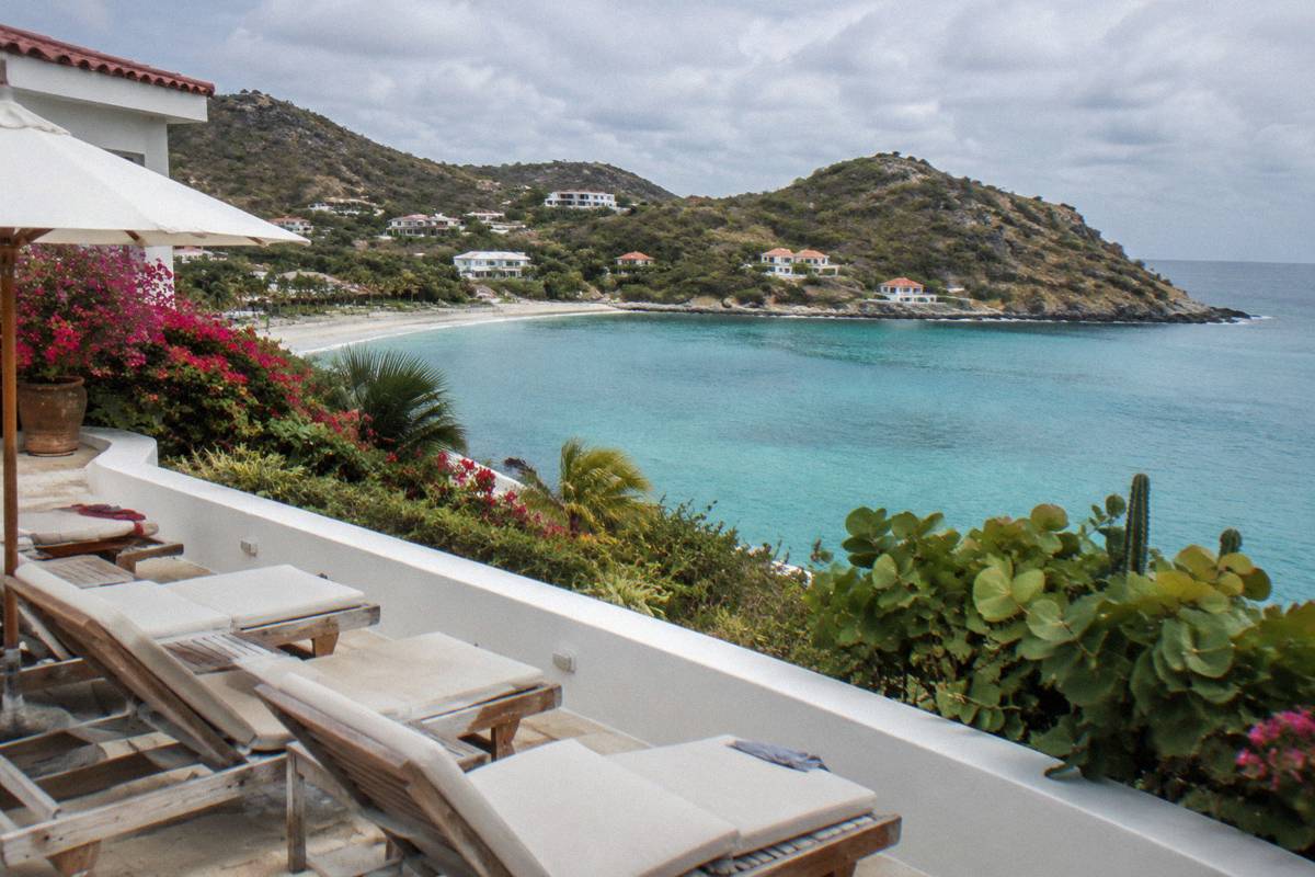 White beachfront hotel terrace with sun loungers facing a calm bay and wooded headland