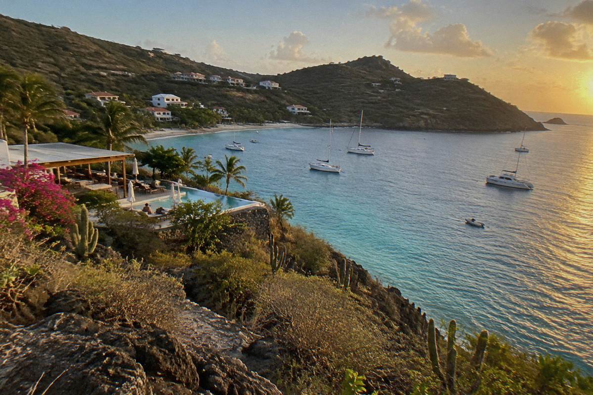 Rocky promontory over a bright bay with a hotel terrace and small boats anchored offshore