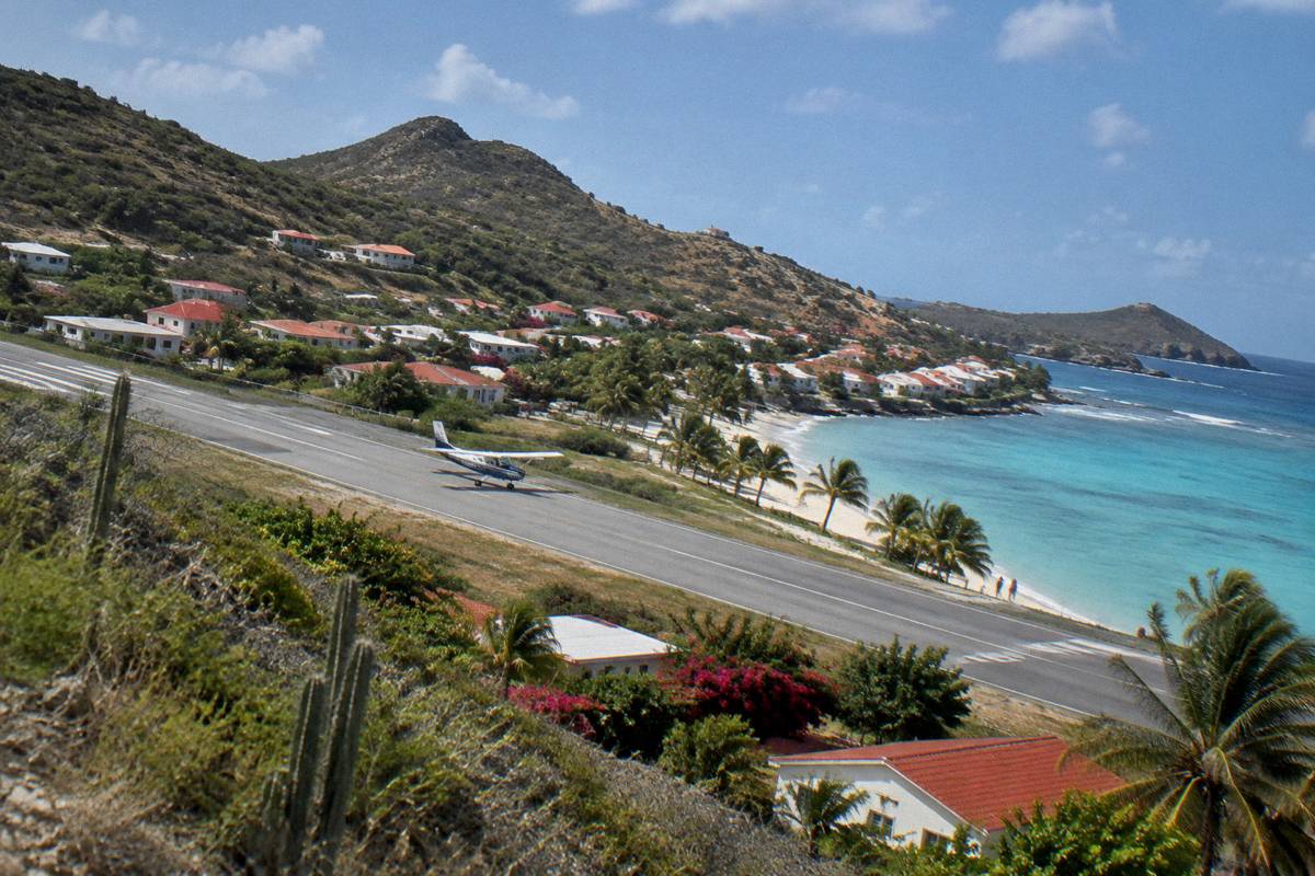 Small aircraft on a short runway beside a palm-lined beach with a village behind