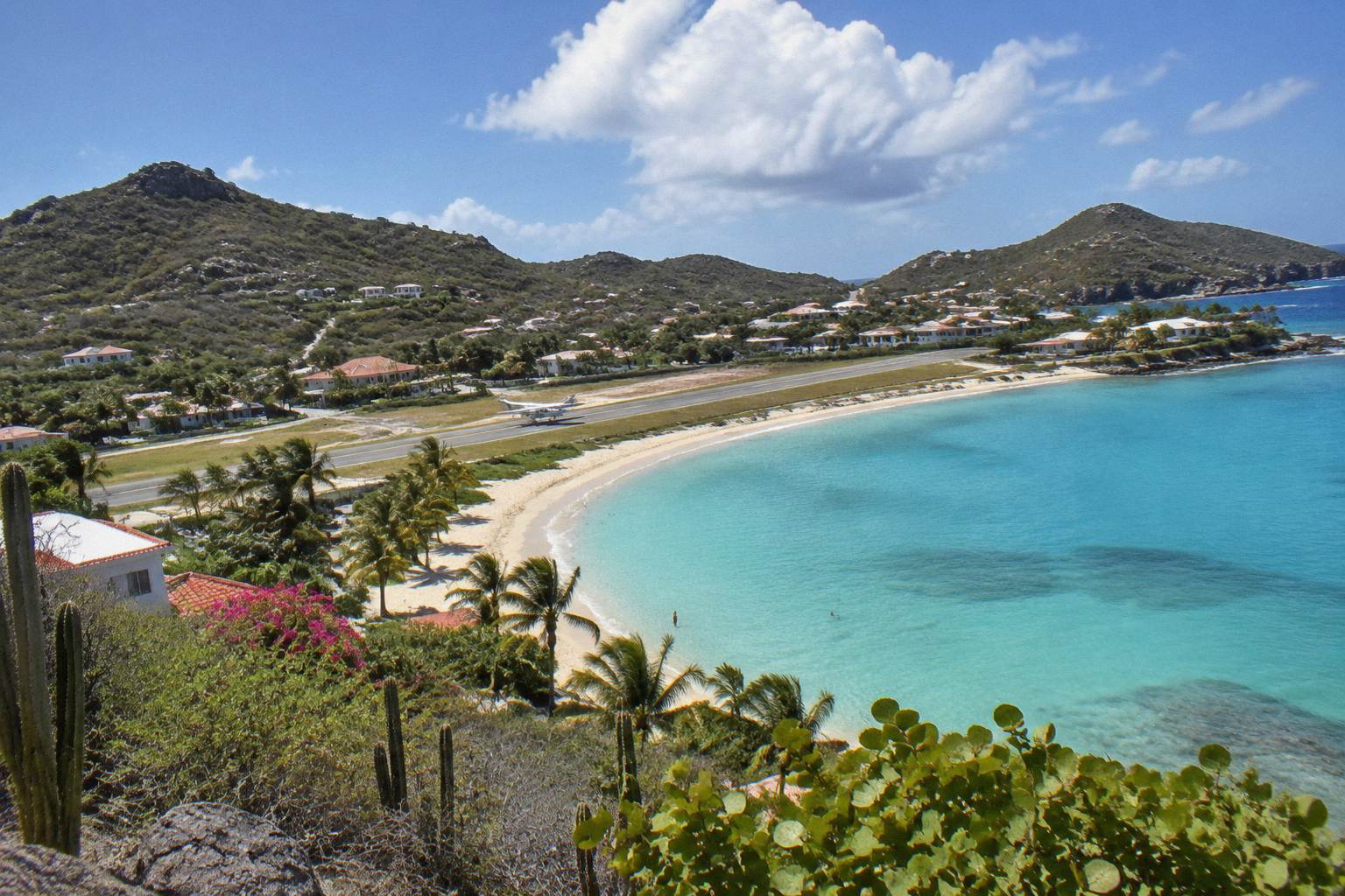 Turquoise bay with a curve of sand, low buildings among palms and a small aircraft on a runway beside the beach