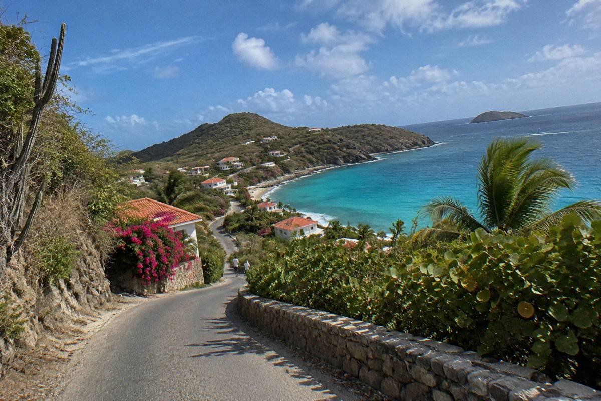A narrow island road curving past tropical vegetation toward a cluster of rooftops near the sea