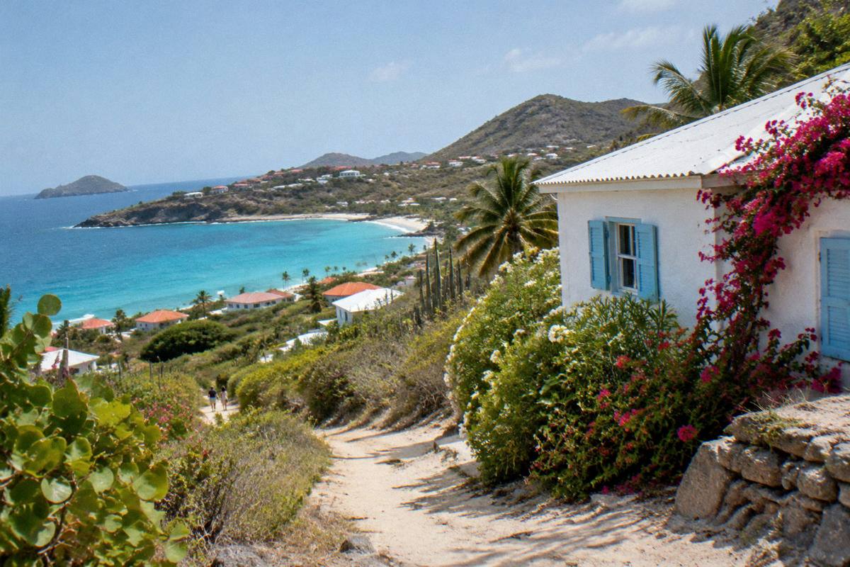 A small whitewashed rental house with blue shutters and flowering bushes beside a sandy path