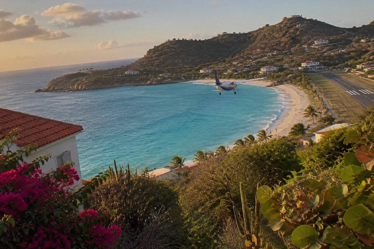 A turquoise bay with a small propeller aircraft descending toward a runway beside the beach