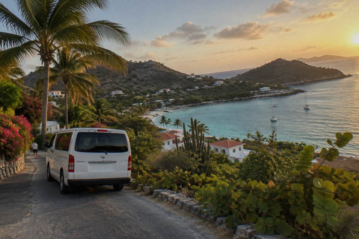 A white taxi van parked on a narrow road beside palm trees and a hillside village