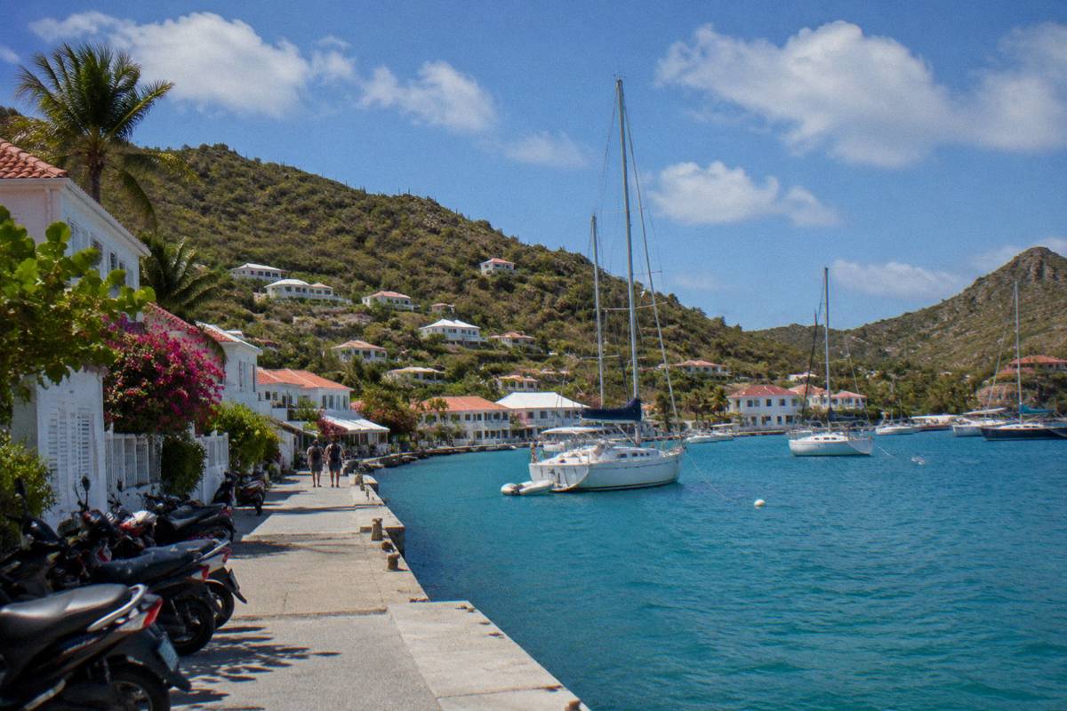 A harbour front with white buildings, parked scooters and moored sailboats at midday