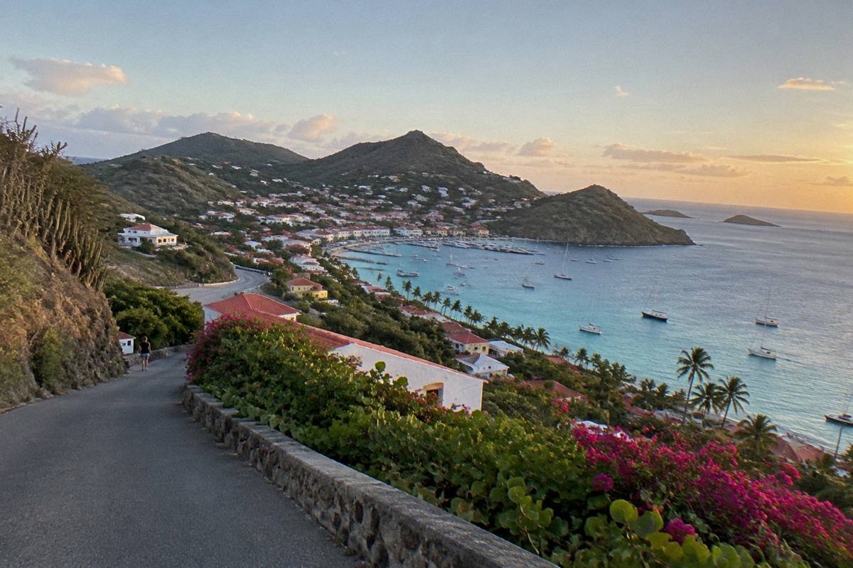 A hillside road at dusk overlooking a lit harbour town and calm sea