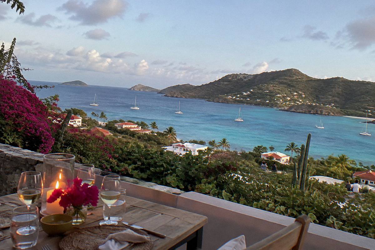 A candlelit dinner table on a terrace above a dark sea with distant anchor lights
