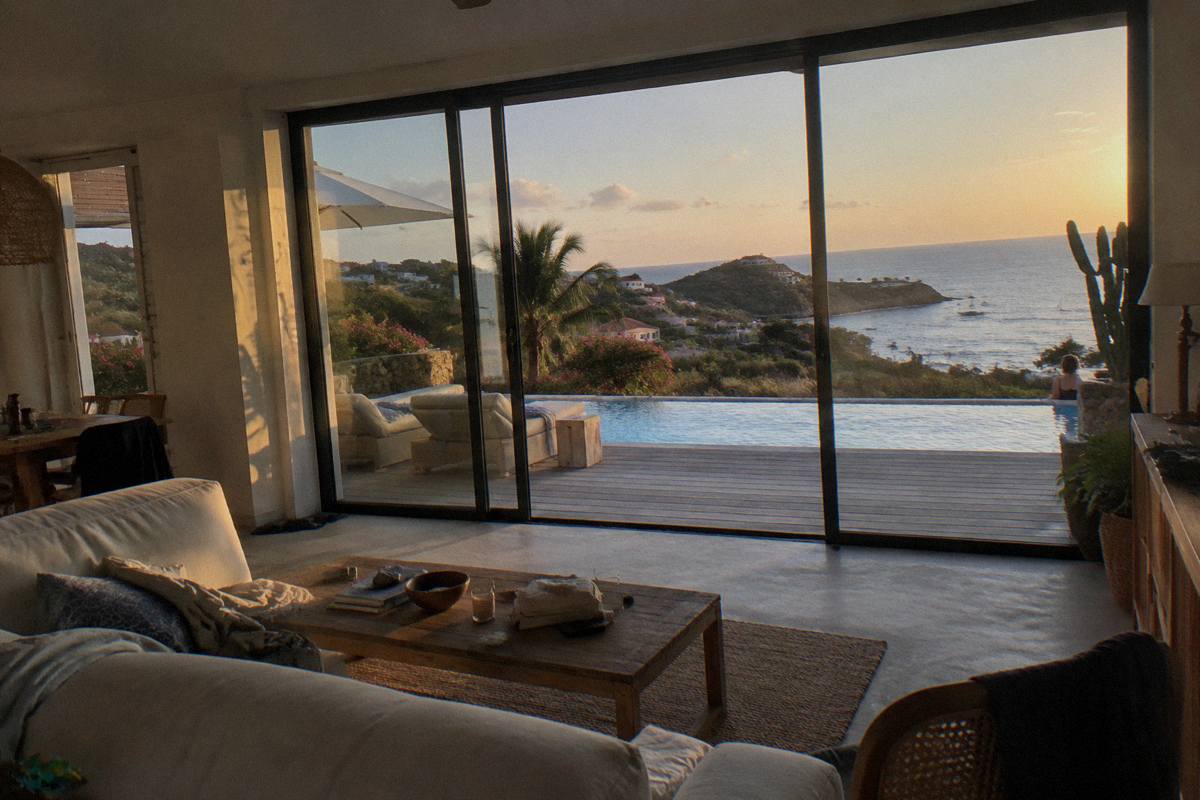 A modern villa living room with floor-to-ceiling glass doors opening onto a pool deck and the sea beyond