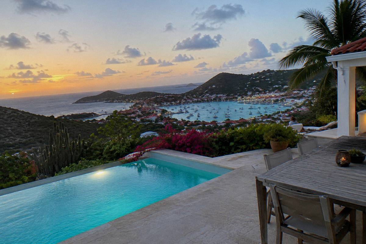 A wide villa terrace at twilight with a lit pool, outdoor dining table and the harbour lights of a town in the distance
