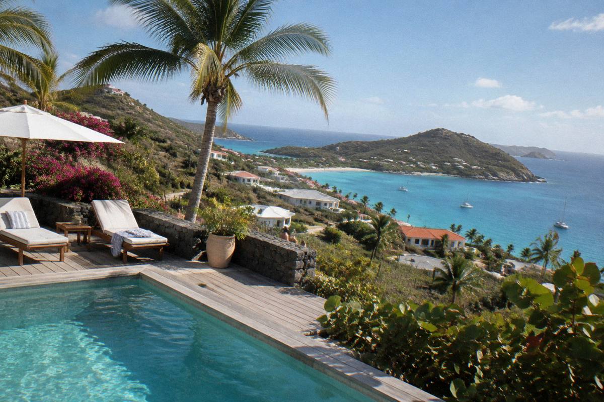 A villa pool deck in morning light with palm trees, sun loungers and a calm sea in the background