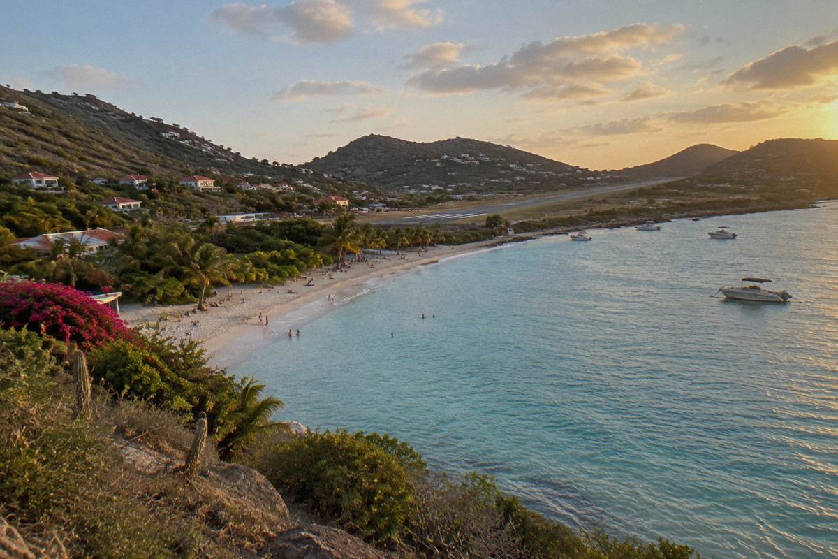 A sandy beach along a busy bay with a small airstrip beyond the palms and hills in the background