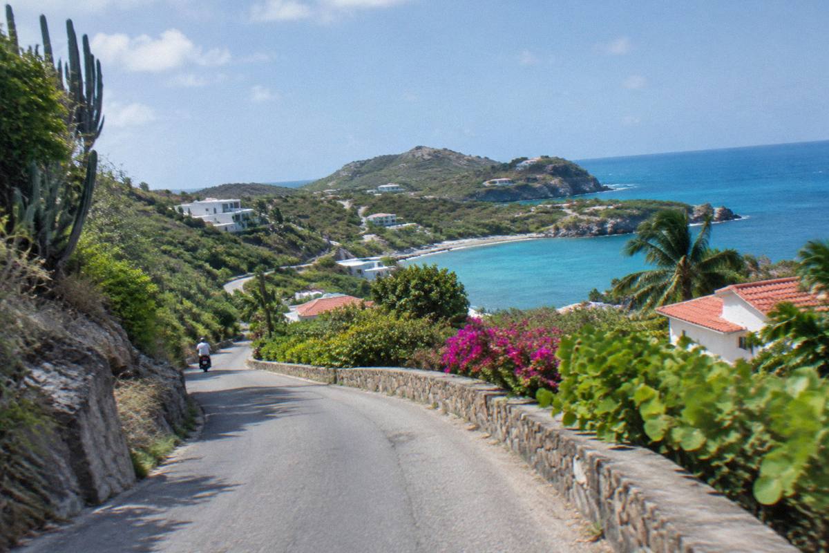 A narrow winding island road climbing between low green hills with a glimpse of the sea